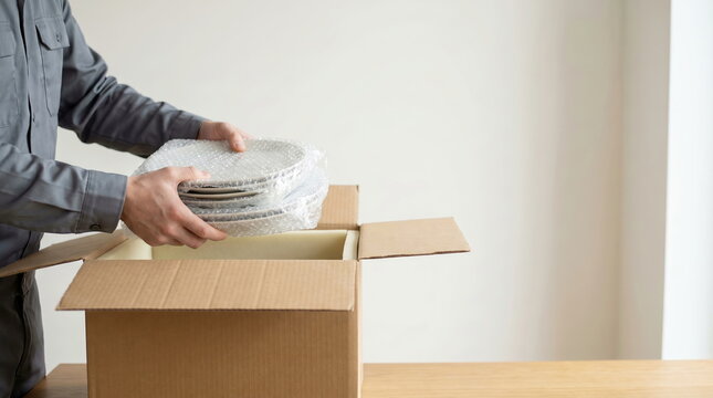 Person packing fragile dishes into cardboard box with bubble wrap, illustrating safe transport during relocation.