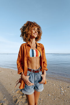 Young woman with curly hair smiles happily standing on the beach wearing a colorful top and denim shorts.