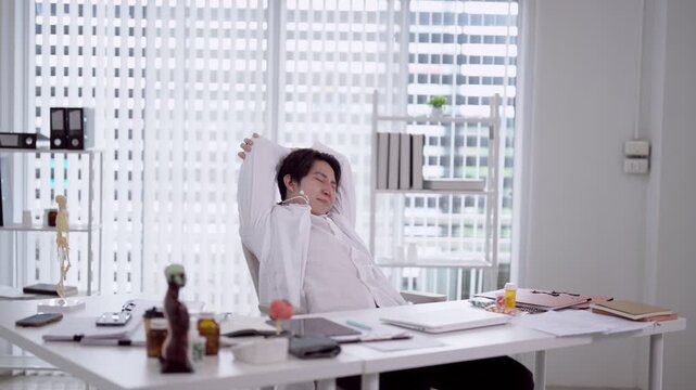 Young Asian male doctor stretching arms overhead during work break in bright modern medical office