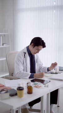 Young Asian physician examining medication capsules while writing prescription notes at modern clinic desk with copy space