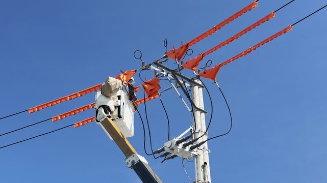 Electrical line worker operating in bucket truck installing orange protective line guards on high voltage power lines. Electrician repairing utility pole grid system under clear blue sky outdoor.