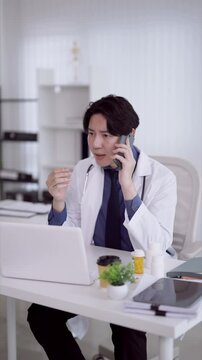 Asian male doctor discussing patient care on phone while gesturing at laptop in bright clinic office