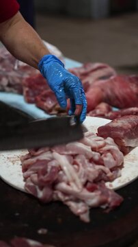 Close up of butcher hands in blue gloves cutting raw fresh pork meat with sharp cleaver knife on round wooden board. Vendor slicing meat pieces at local outdoor market counter, food store concept.