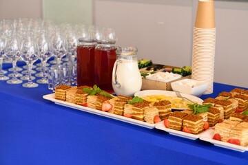 Catering coffee break buffet table with slices of honey cake, milk glass jug, red berry mors drink, empty wine glass rows and paper cup stack. © Tatiana
