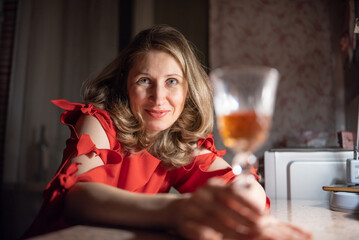 Thoughtful caucasian woman in red dress sitting at a bar or cafe at night, holding a smartphone, with a blurred glass of wine or cocktail in the foreground with dim lighting. © shymar27