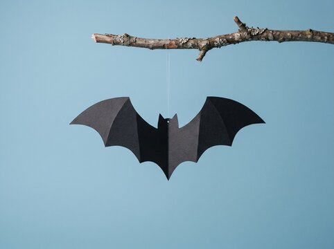 A black paper cutout bat flying under a dry wooden branch against a plain light blue background surface