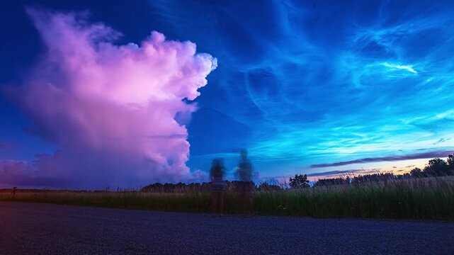 View of two people standing on a road at dusk observing a dramatic sky filled with vibrant noctilucent clouds and a large pink cloud formation in Riga, Latvia.