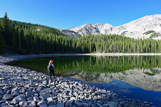 Woman explores shore of second Thomas Lake in Maroon Bells-Snowmass Wilderness in White River National Forest on calm clear summer morning.