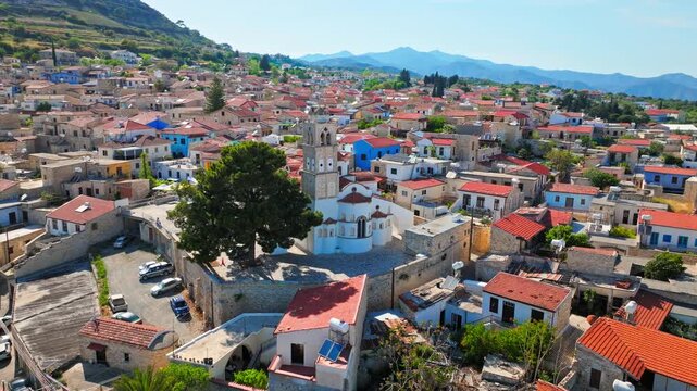 Drone view of Pano Lefkara village square and rooftops in Cyprus. Traditional houses, trees and distant mountains create a scenic countryside view.
