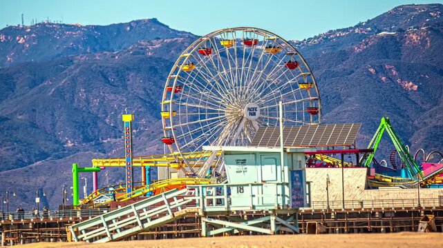 View of the iconic Pacific Park ferris wheel and amusement rides on Santa Monica Pier with mountains in the background in Santa Monica, California, United States.