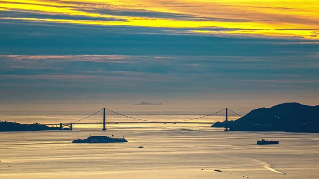View of the Golden Gate Bridge at sunset with a cargo ship passing near Alcatraz Island under a vibrant sky in San Francisco, California, United States.
