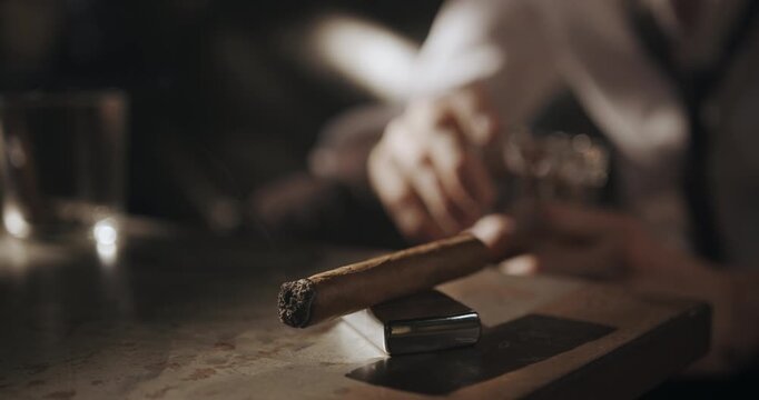 Man picks up a glass of whiskey beside a smoking cigar, puts the drink back and reaches for the cigar.