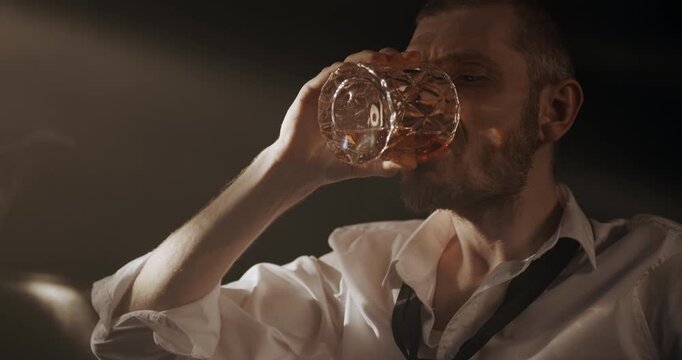 Tired bearded man drinks whiskey from a glass and leans back on a leather sofa in a dim room.