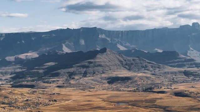 Aerial view of the rugged Drakensberg mountain range rising above vast golden plains and rural settlements in Thabo Mofutsanyana District Municipality, Free State, South Africa.