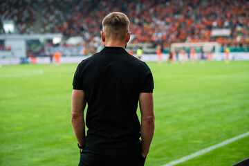Soccer coach standing on the sideline and watching a football match in a modern stadium, with green field and spectators in background. © Dziurek