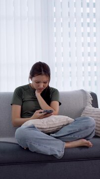 Young Asian woman with upset expression scrolling smartphone while sitting cross-legged on living room sofa with copy space