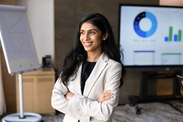 Obraz premium Portrait of Indian businesswoman or corporate trainer standing with crossed arms in corporate office during business presentation, financial charts and graphs displayed on digital screen in background