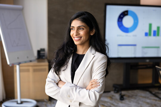 Portrait of Indian businesswoman or corporate trainer standing with crossed arms in corporate office during business presentation, financial charts and graphs displayed on digital screen in background