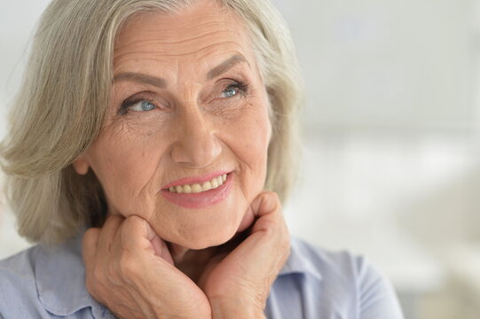 elderly woman smiling with hands on chin in a close-up, front view of an indoor setting with neutral walls and light skin tone, conveying a content mood, ideal.