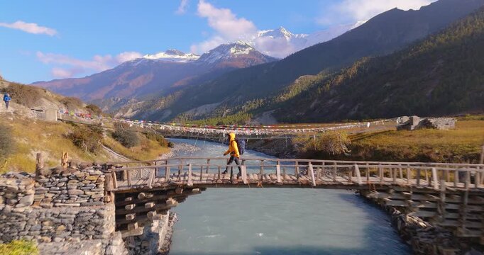 Annapurna Circuit, Manang Nepal, Drone pushes forward towards a shot of a male hiker in a yellow jacket crossing a wooden bridge over the Marsyangdi River with snow-capped mountains in the background.