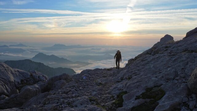 A lone hiker with a backpack on a rocky mountain trail in Spain's Picos de Europa. Breathtaking sunset over a majestic cloud inversion and rugged peaks creates a serene, remote landscape.