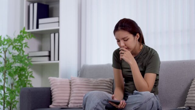 Frustrated young Asian woman feeling anxious while using smartphone on couch in modern living room with copy space
