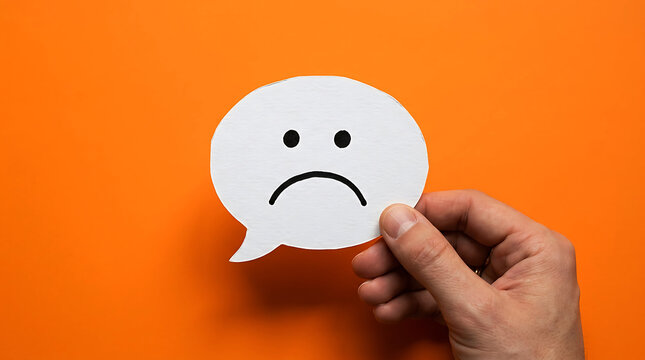Hand holding a white speech bubble with a sad face icon on an orange background