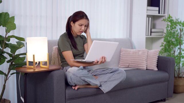 Young Asian woman feeling stressed and frustrated working on laptop sitting cross legged on sofa at home
