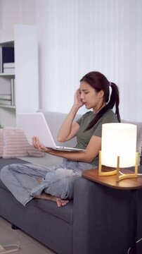 Stressed young Asian woman holding laptop while sitting on sofa in modern apartment with copy space