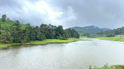 Obraz premium Scenic mountain lake surrounded by lush green hills, forest trees and misty clouds under overcast sky, capturing peaceful monsoon landscape, nature travel and hill station countryside view.