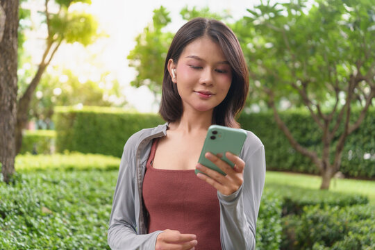 Asian woman using smartphone fitness tracking app checking AI fitness application on smartphone in green urban park wellness and healthy lifestyle.
