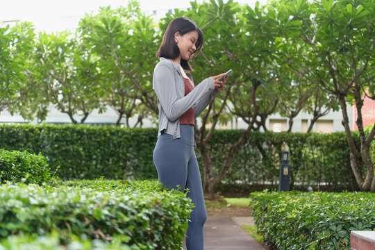 Asian woman using smartphone fitness tracking app checking AI fitness application on smartphone in green urban park wellness and healthy lifestyle.