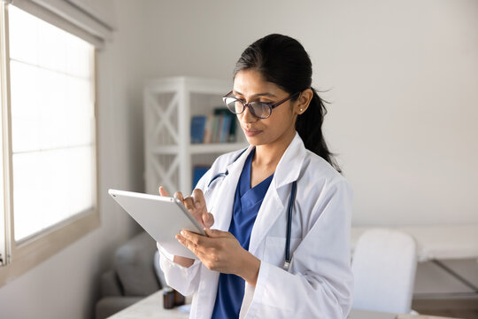 Focused South Asian female doctor working in clinic office using digital tablet, reviewing electronic health records, checking patient data, updating medical report. Healthcare technology at workplace