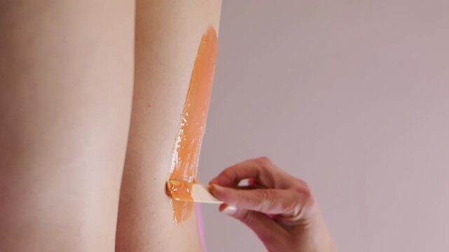 Vertical video. Body care. Healthy treatment. Unrecognizable professional beautician hands waxing legs of woman with orange sugaring paste using spatula in spa salon.