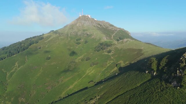 Aerial view pulling back from the impressive mountain La Rhune in Nouvelle-Aquitaine, France. The drone reveals vast, green slopes and a communication complex on the peak under a sunny sky.