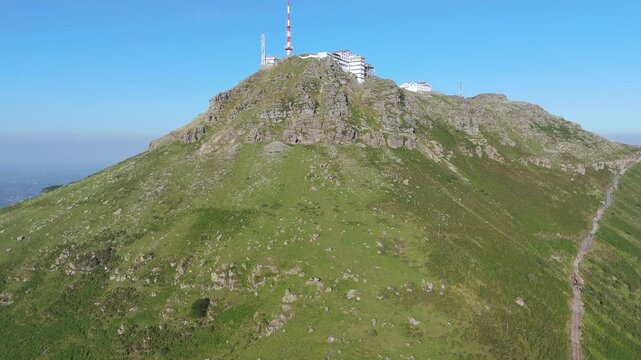 Aerial drone pull back shot revealing the La Rhune mountain peak with its radio tower and observatory in the Pyrenees, Nouvelle-Aquitaine, France. A lush, rocky landscape under a clear blue sky.