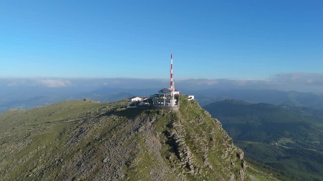 Aerial pan left and tracking shot over La Rhune viewpoint on mountain in, France. Majestic Pyrenees landscape with a telecommunication facility on the rocky summit.