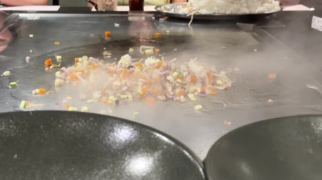 Chef stir fries oriental style rice with vegetables on steaming hot griddle cutting it with serving spatula in slow motion