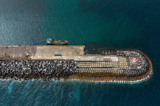 Aerial view of a harbor breakwater with a cargo ship docked alongside, protected by concrete tetrapods and featuring a small lighthouse at the end in Madalena, Azores, Portugal.