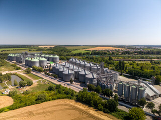 High angle view shows large industrial grain elevator facility with metallic silos near green fields under clear blue sky. © Pasha