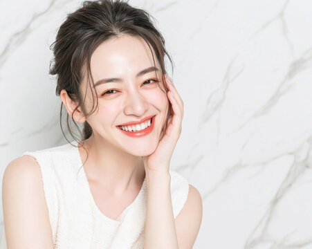 Portrait of beautiful young East Asian woman resting chin on hand with radiant smile showing white teeth against white marble background for beauty skincare cosmetics concept