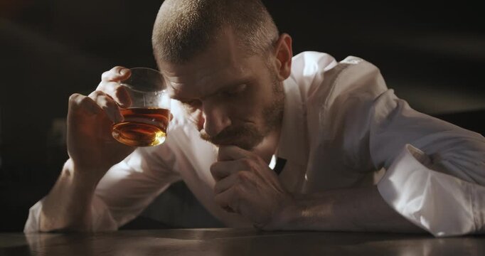 An exhausted man leans over the table holding a glass of whiskey, looking down with a weary and defeated expression.