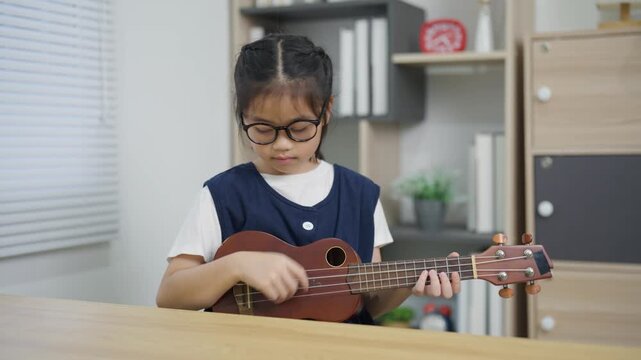 Young girl wearing glasses playing ukulele at music room with acoustic guitar electric guitar keyboard and amplifier background