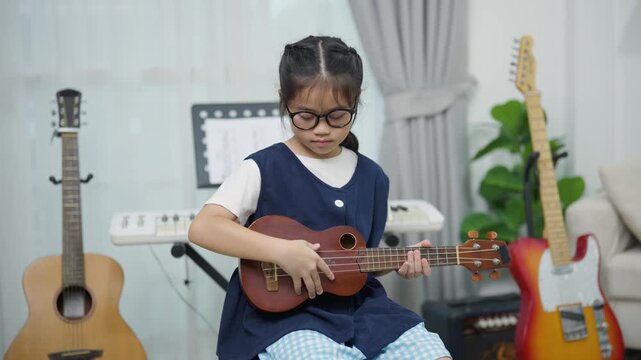 Young girl wearing glasses playing ukulele at music room with acoustic guitar electric guitar keyboard and amplifier background