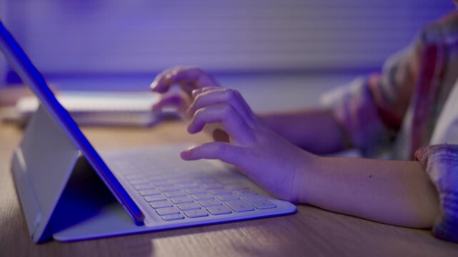 Close up of hands typing on keyboard attached to tablet device on wooden desk with blurred workspace background in evening light