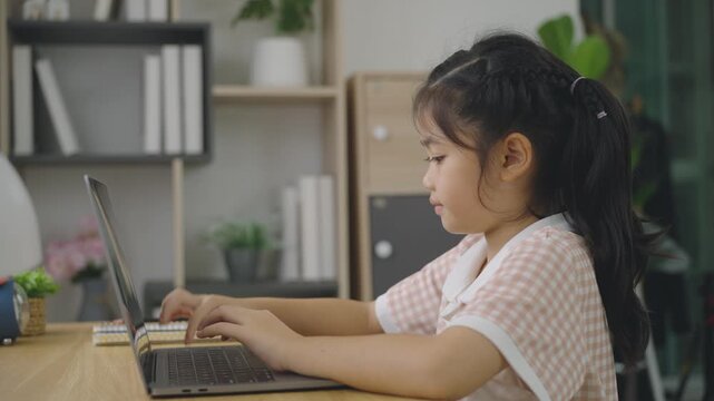 Young girl in gingham dress sitting at wooden desk using laptop computer in cozy living room with shelves and plants behind