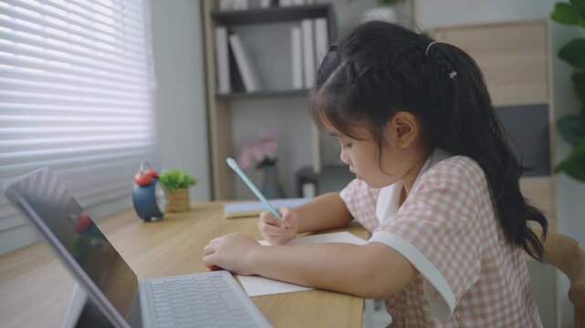 Young girl focused on writing with pencil paper at wooden desk using laptop in bright cozy room with natural light