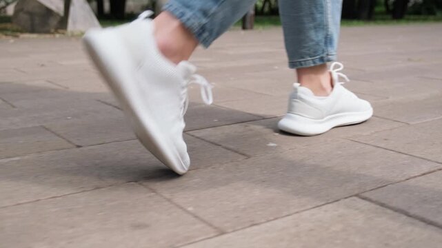 Person strolling outdoors in comfortable white footwear. Low angle shot of a person wearing casual clothes and white running sneakers walking on a city sidewalk. Casual active lifestyle concept.