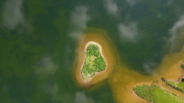 Aerial view of a small, lush green island surrounded by calm, dark water with reflections of clouds, Aldwincle, England, United Kingdom.