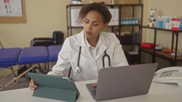 Woman doctor with stethoscope typing on laptop and using tablet at clinic desk in building; concentration compassion care.